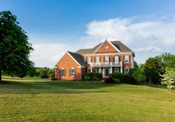 Two-story brick home with a wide green front lawn and mature trees under a blue sky