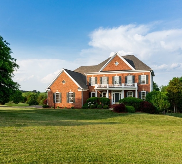 Two-story brick home with a wide green front lawn and mature trees under a blue sky