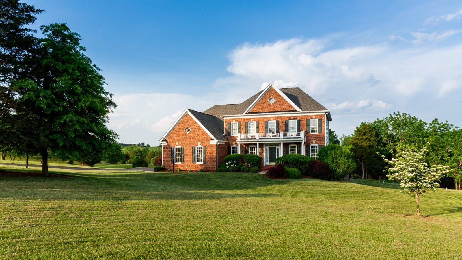Two-story brick home with a wide green front lawn and mature trees under a blue sky